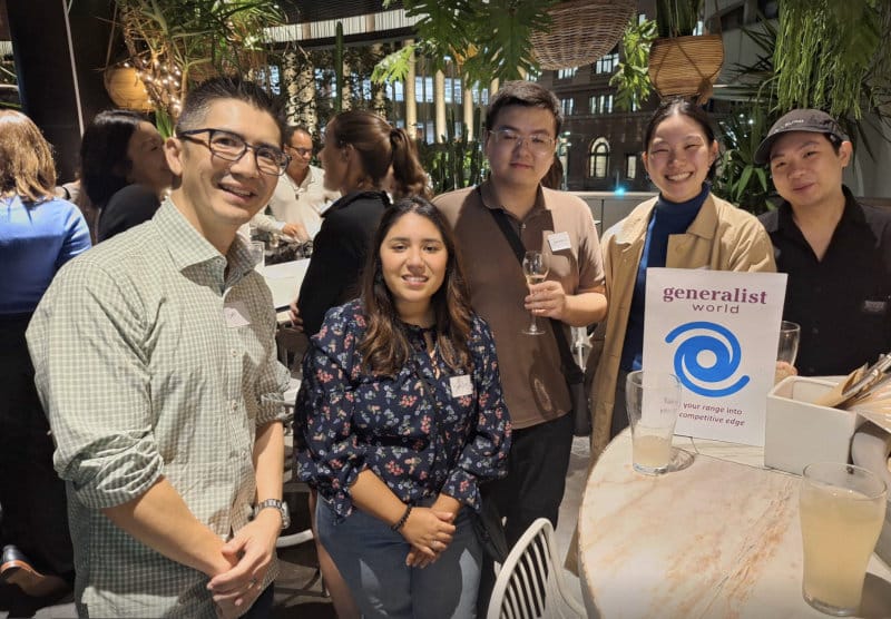 A small group of generalists smiling around a table with a Generalist World sign