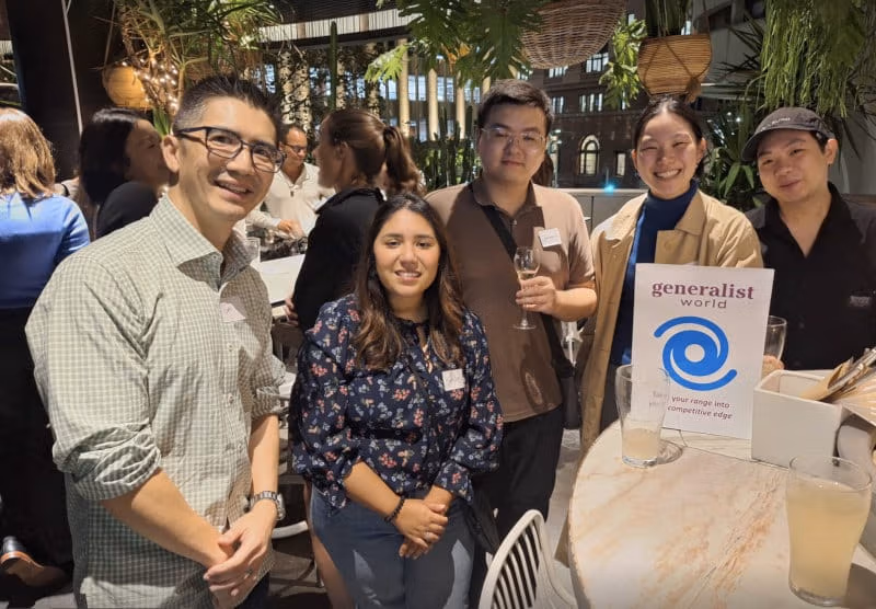 A small group of generalists smiling around a table with a Generalist World sign